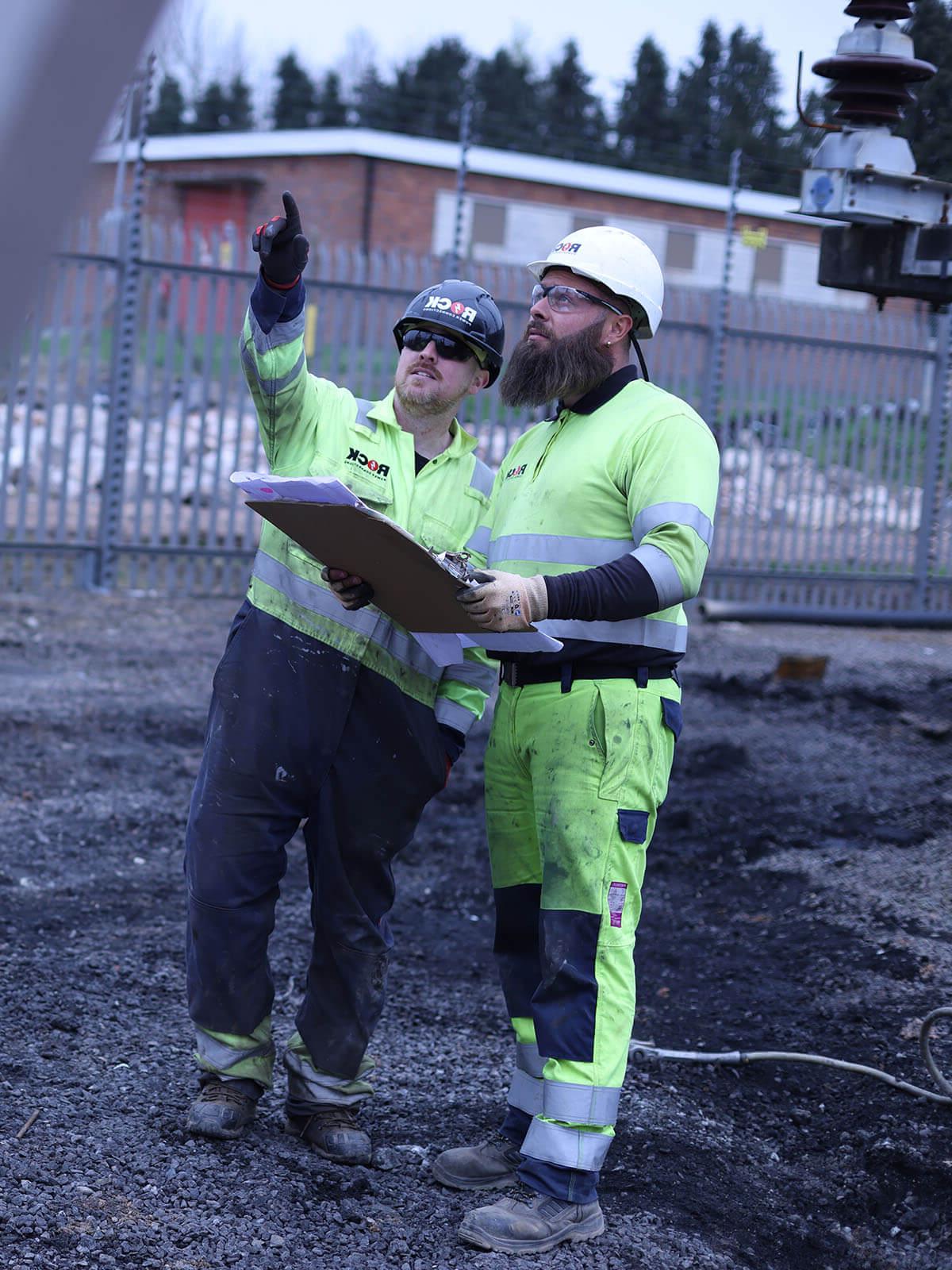 两名男性岩石电力公司员工穿着显眼的衣服, 看着计划，向上指